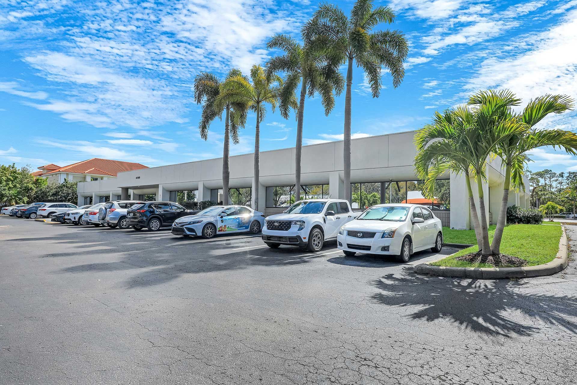 Cars parked in front of a white building with palm trees under a blue sky.