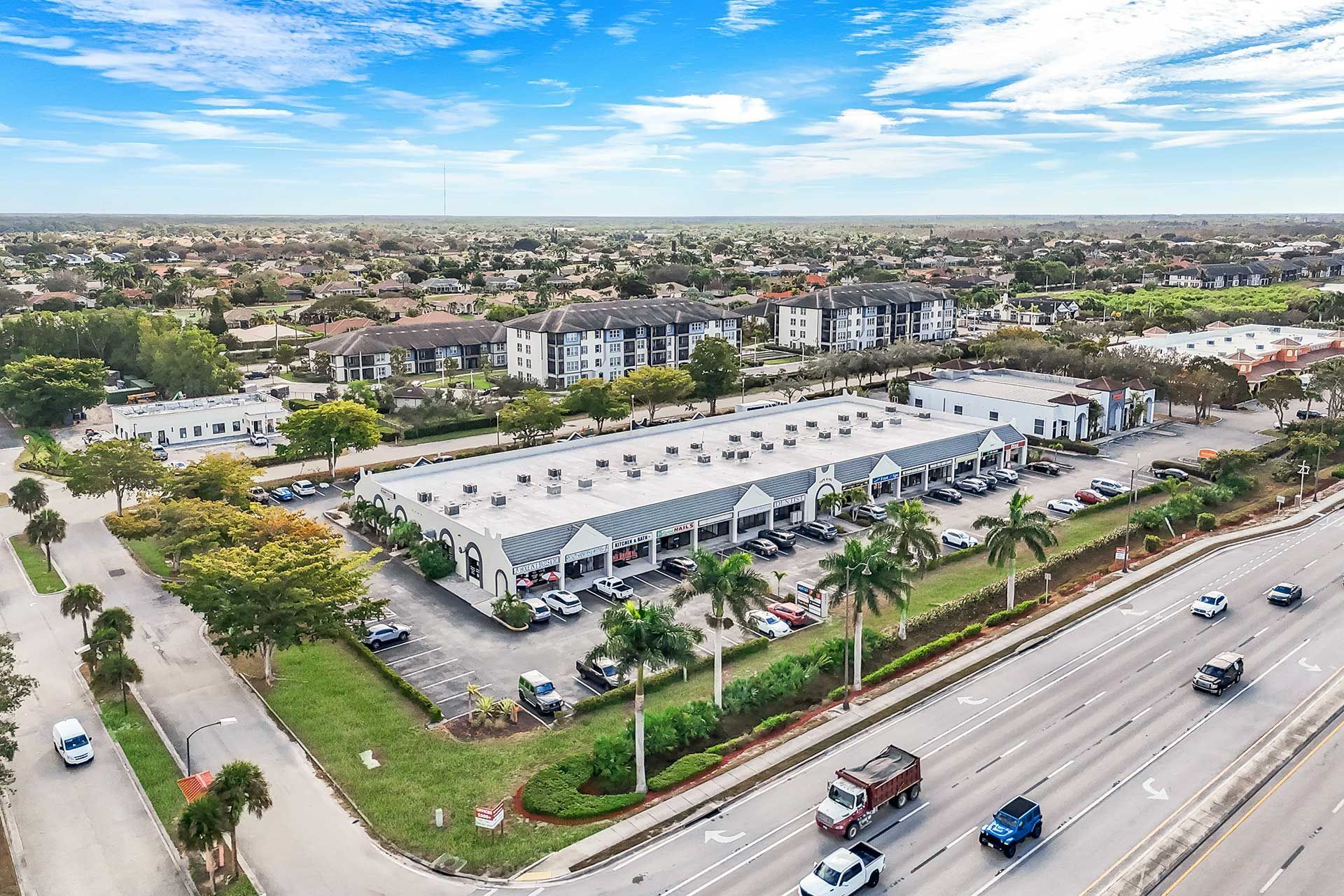 Aerial view of a commercial building with a long, white roof, palm trees, and a road with cars.