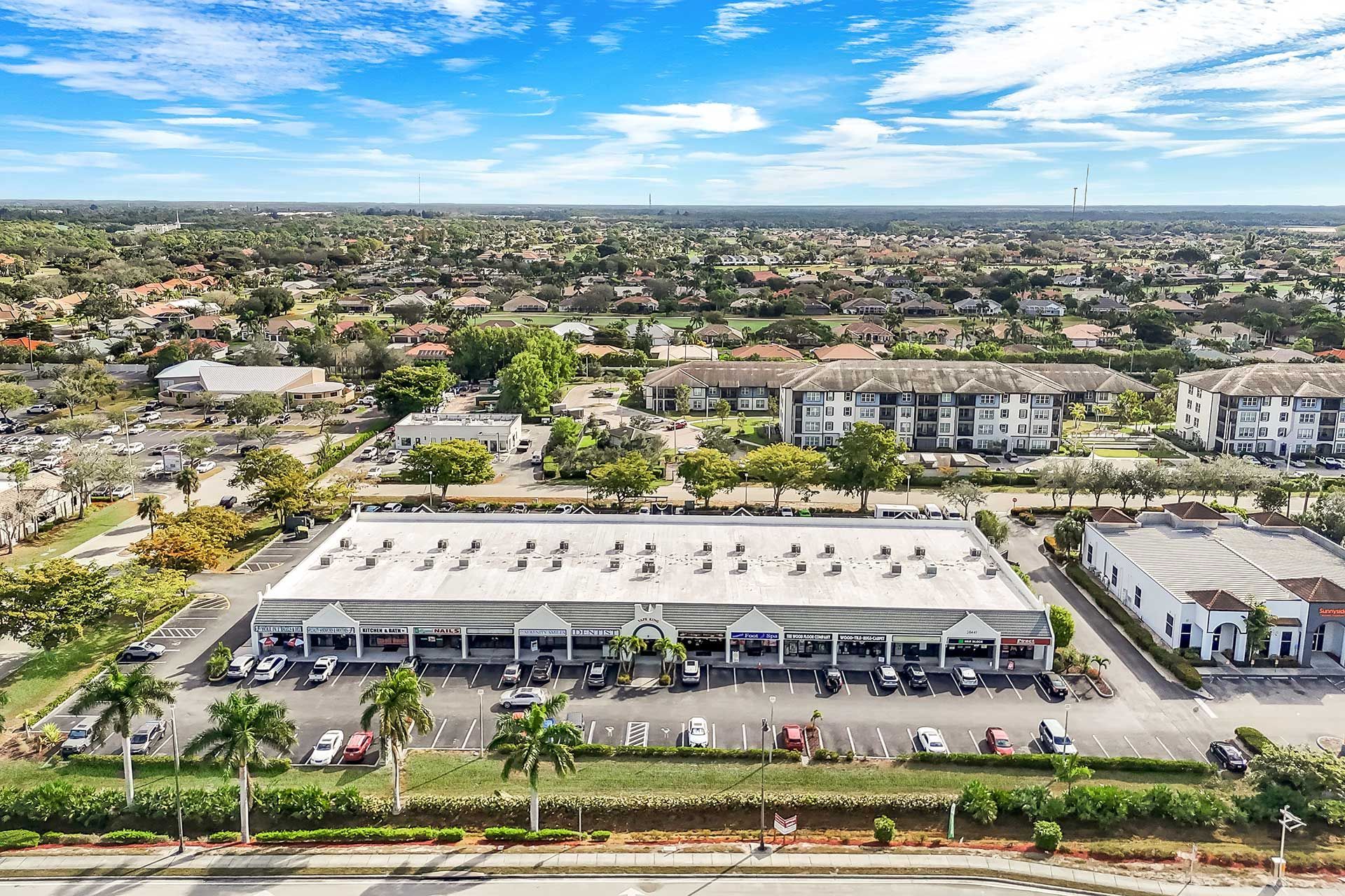 Aerial view of a long, single-story commercial building with parking, trees, and residential buildings in the background.