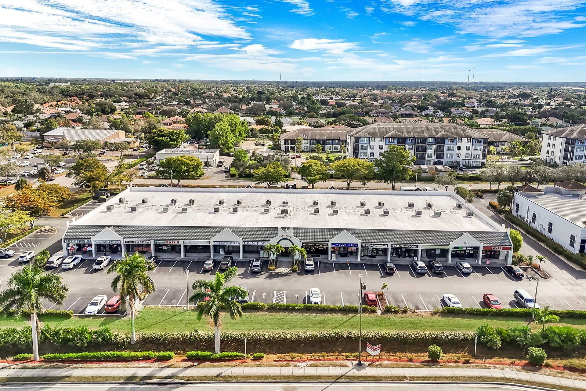 Aerial view of a strip mall with parked cars, surrounded by green lawns, trees, and residential buildings on a sunny day.