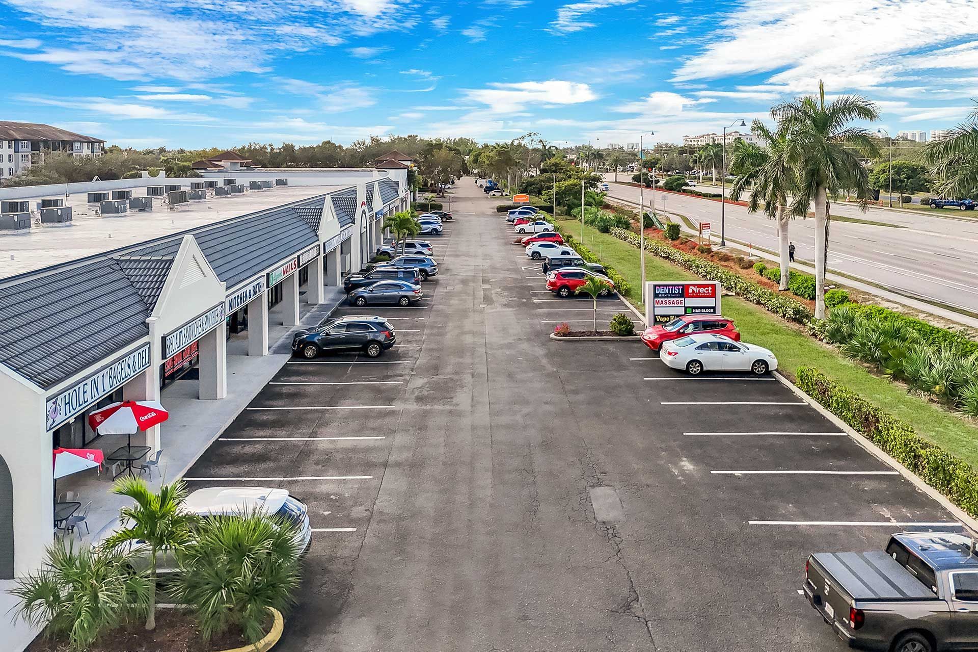 Strip mall with parked cars, palm trees, and storefronts on a sunny day.