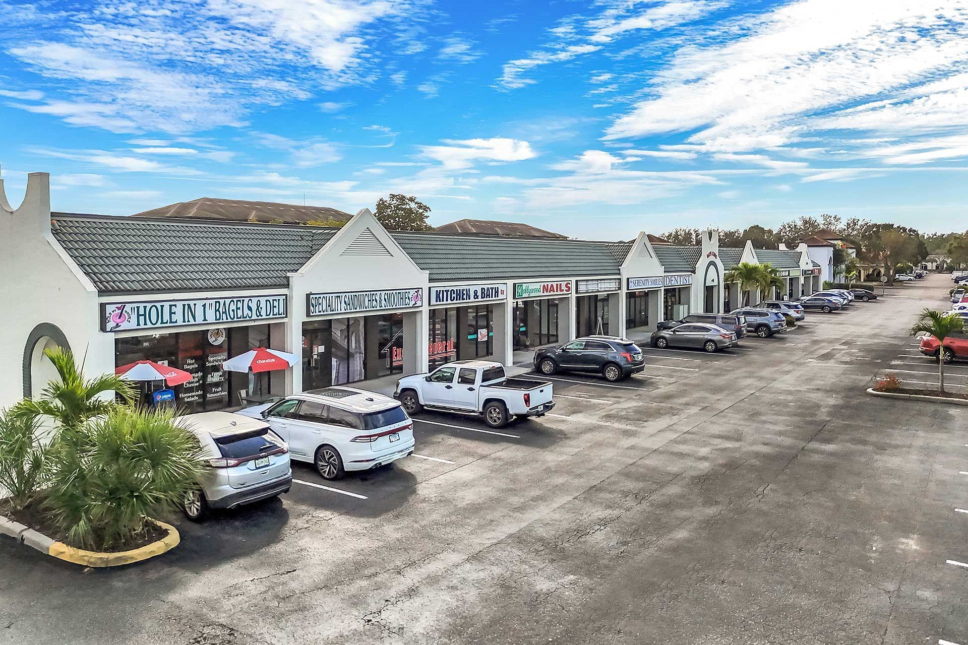 Strip mall with storefronts, parking lot, vehicles, and a blue sky with clouds.
