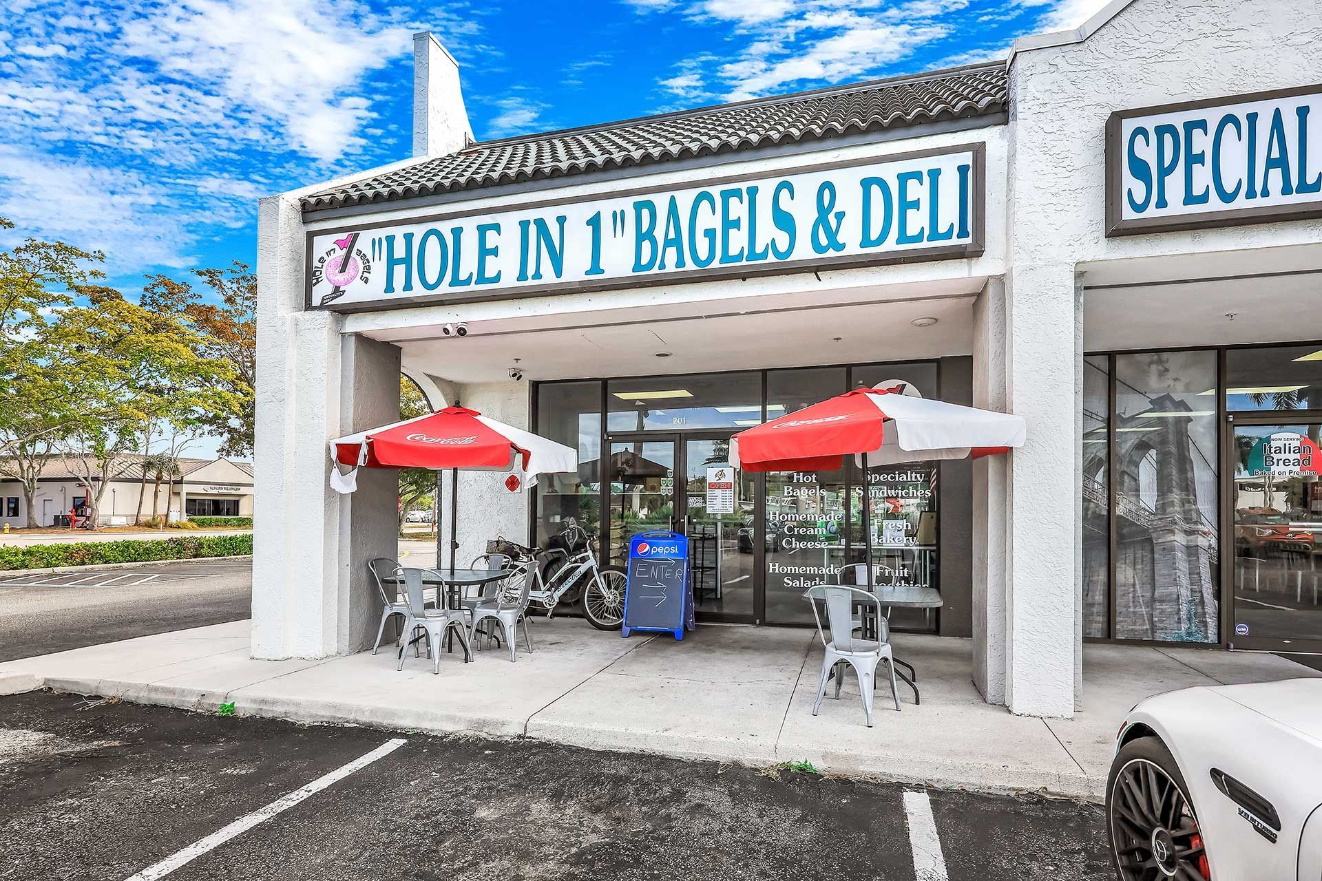 Exterior of Hole in 1 Bagels & Deli with outdoor seating, red umbrellas, and a white building under a cloudy sky.