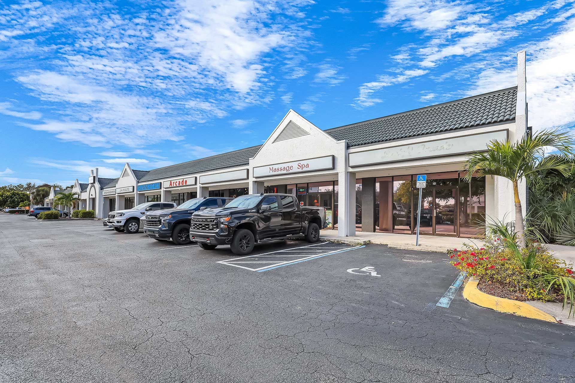 Exterior of a strip mall with a cloudy blue sky. Several cars are parked in front of the businesses.