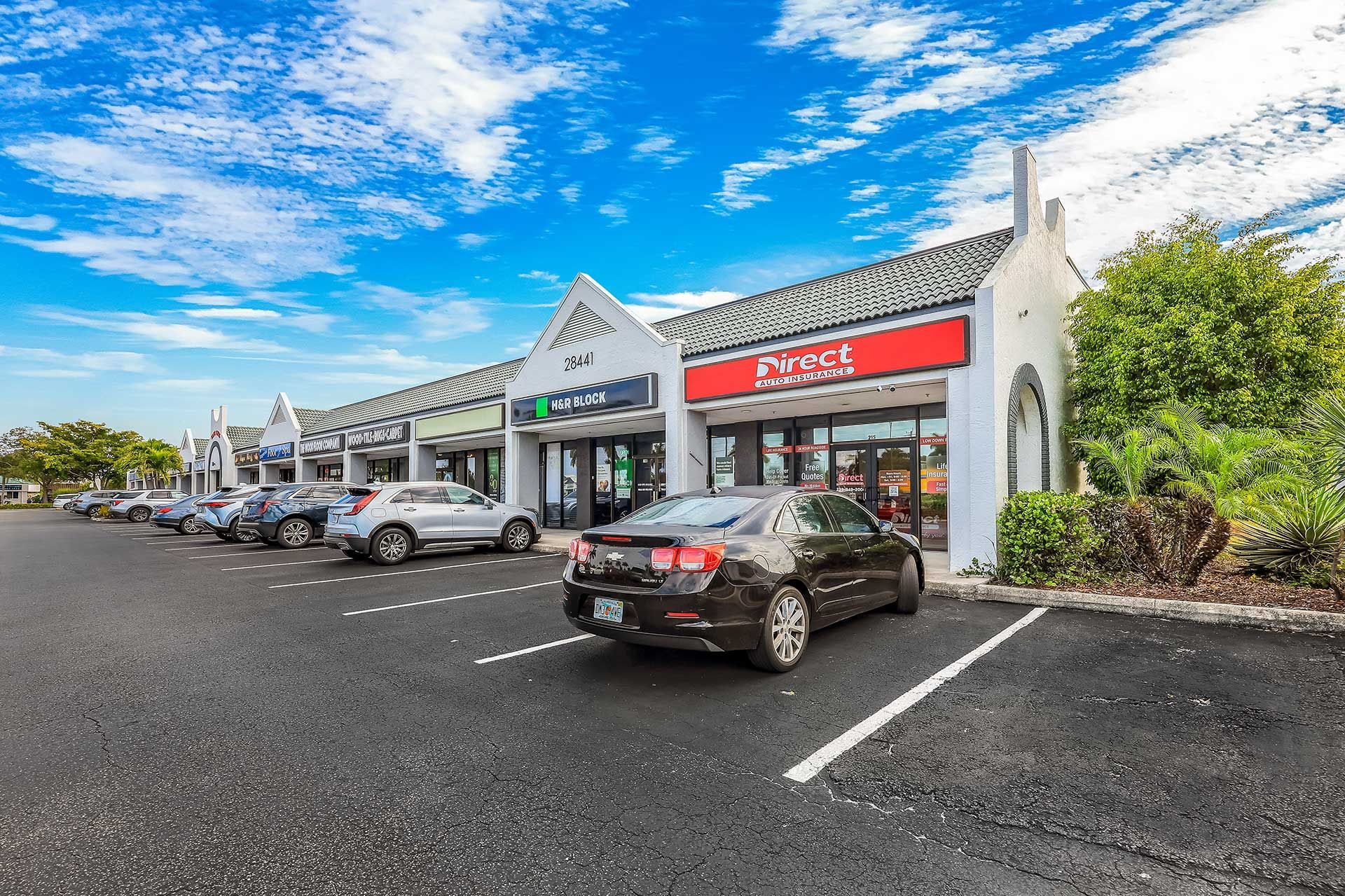 Shopping plaza with parked cars under a bright blue sky. Stores have awnings and red signage.