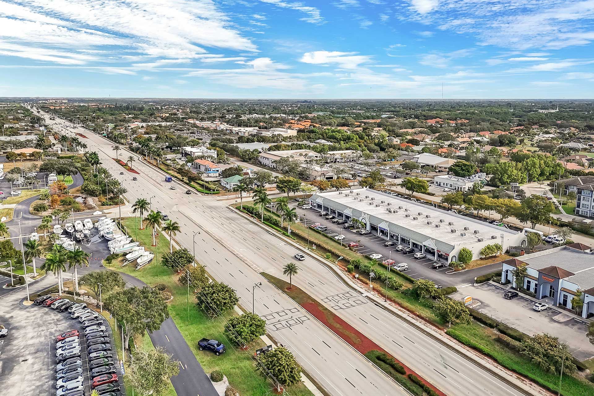 Aerial view of a wide road with buildings and parked cars on both sides under a partly cloudy sky.