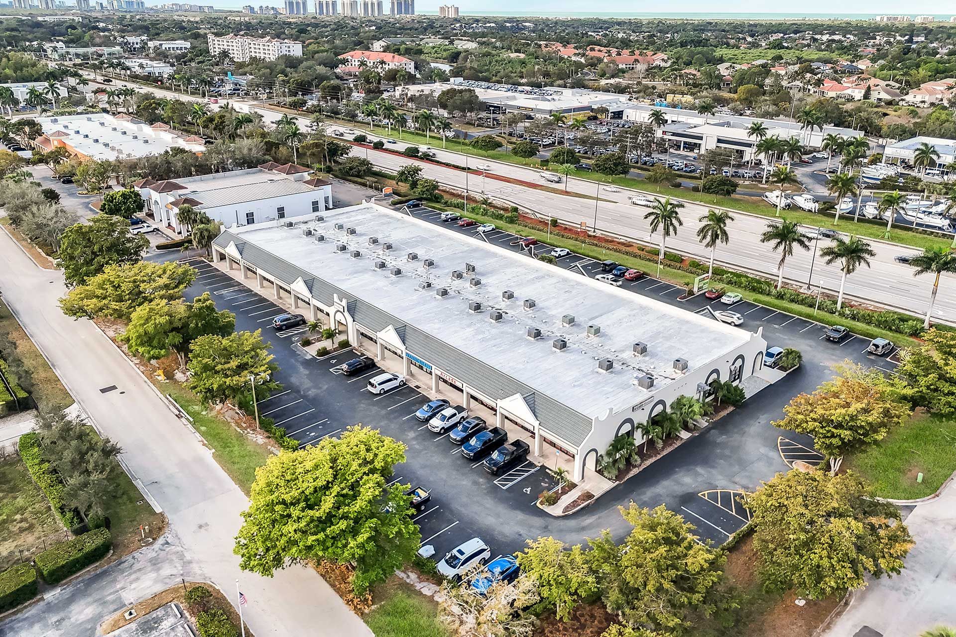Aerial view of a long, gray building with parking lot and surrounding greenery.