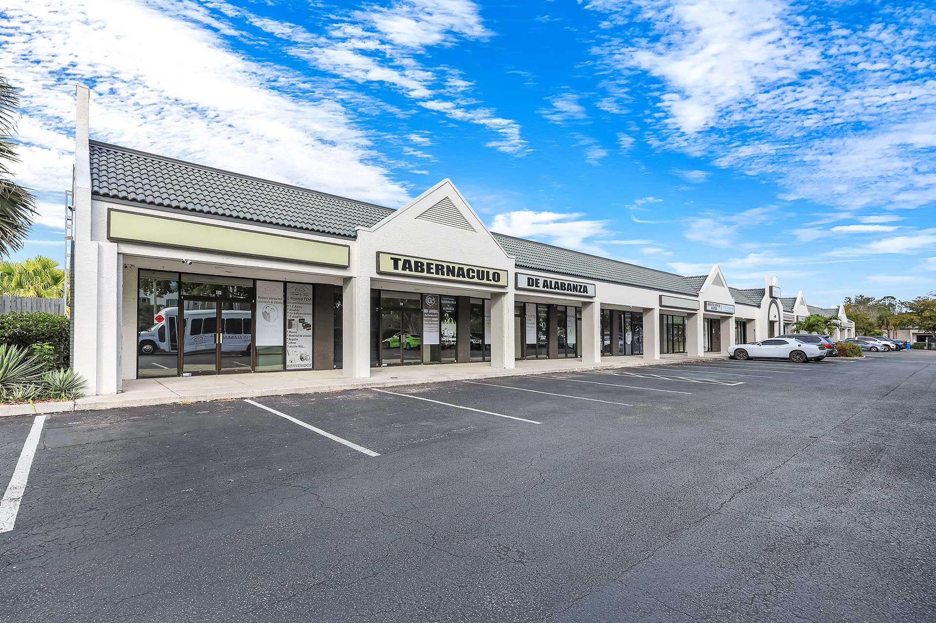 A strip mall with a row of shops with glass storefronts and a parking lot on a sunny day.