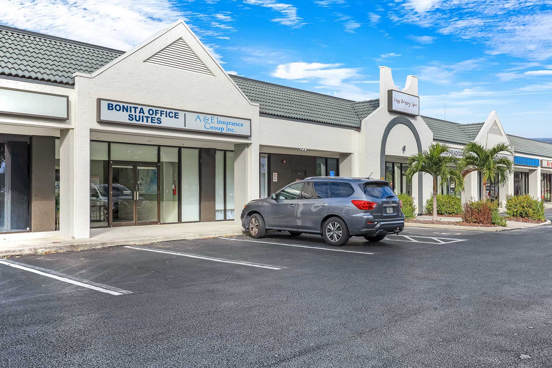Gray SUV parked in front of a white strip mall with storefronts under a blue sky.