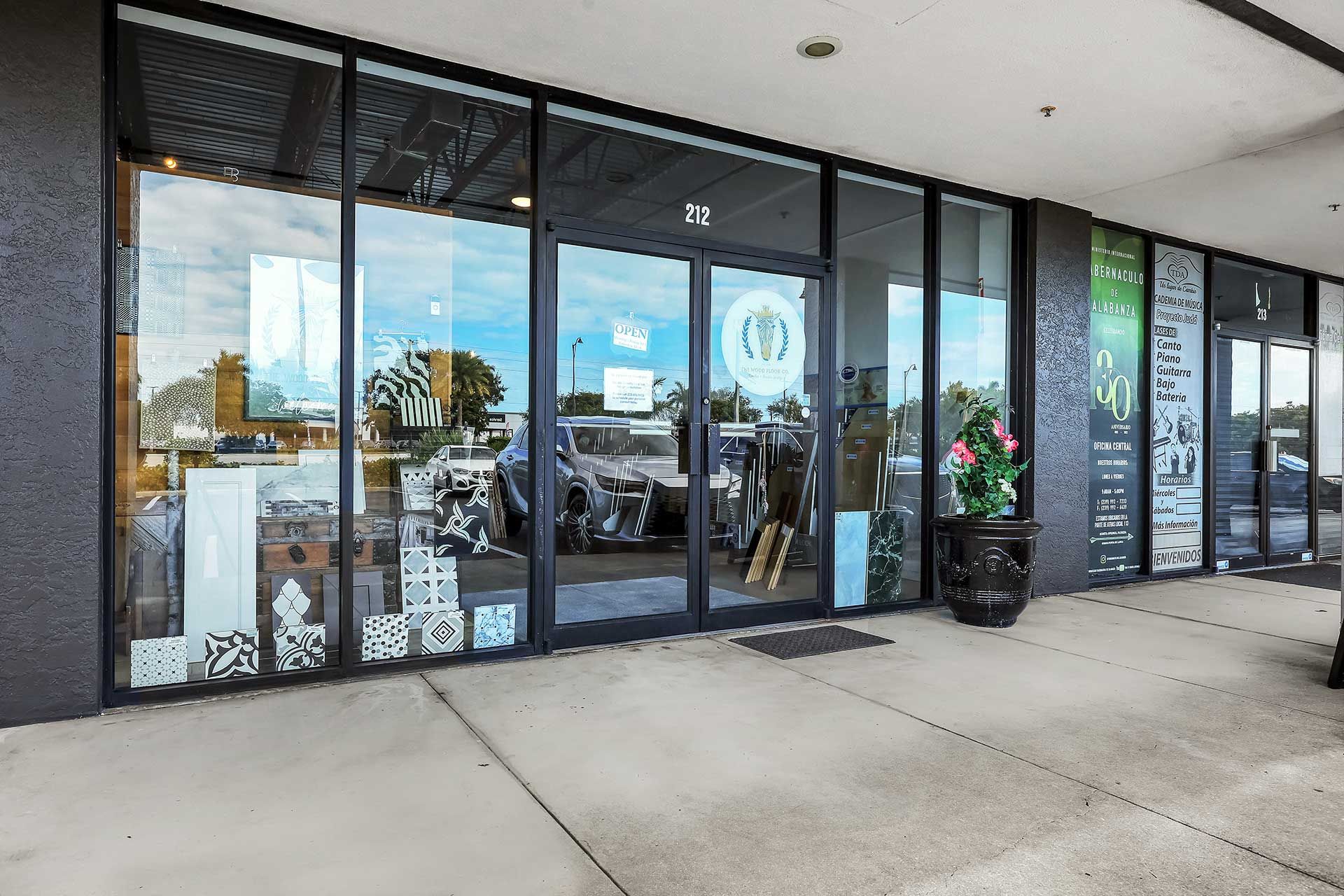 Exterior of a store with large windows and a sign above the door. A black planter with flowers sits outside.