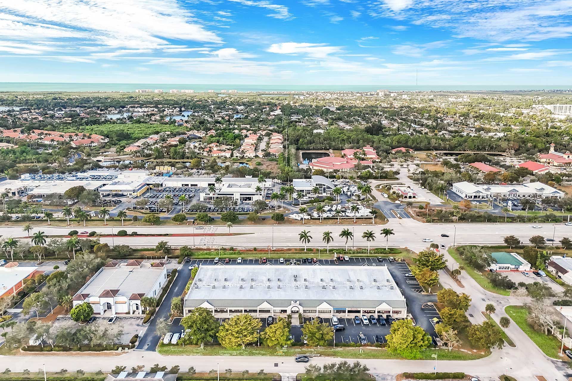 Aerial view of a commercial area with buildings, roads, and a residential area in the background under a blue sky.