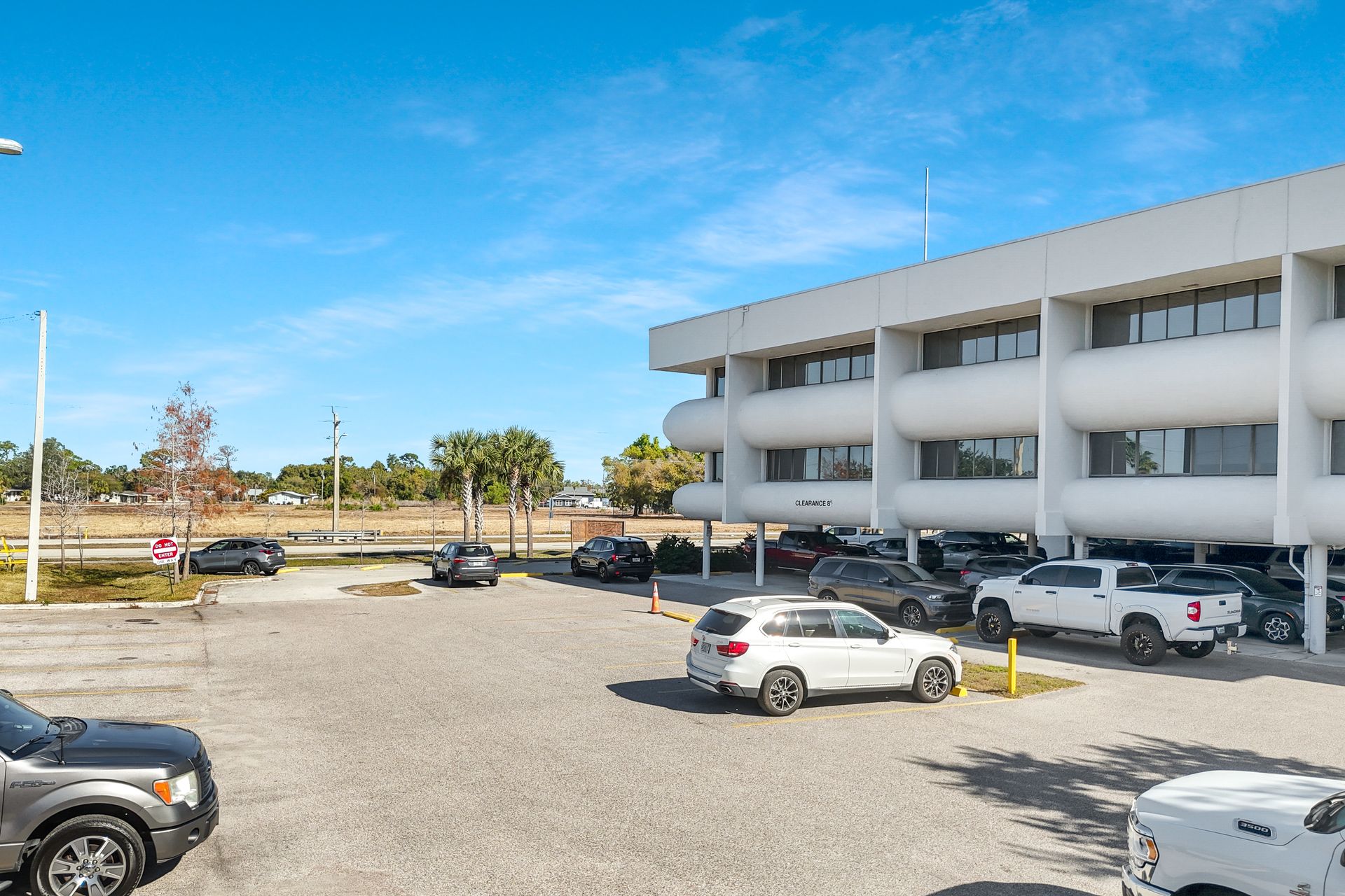 Two-story white building with cars parked in front on a gravel lot under a clear blue sky.