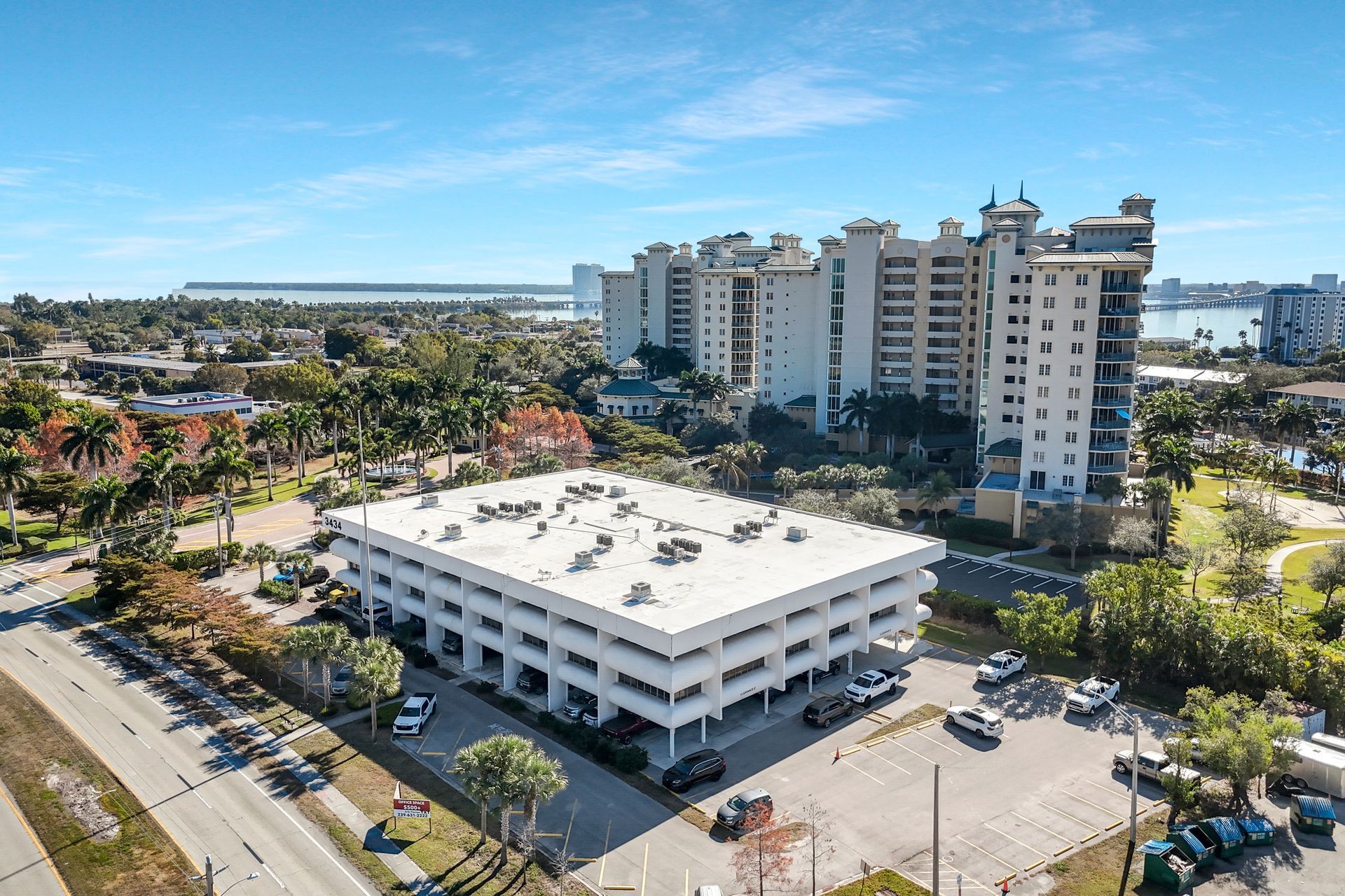 Multi-story white office building with parking lot, residential buildings, and waterway in the distance.
