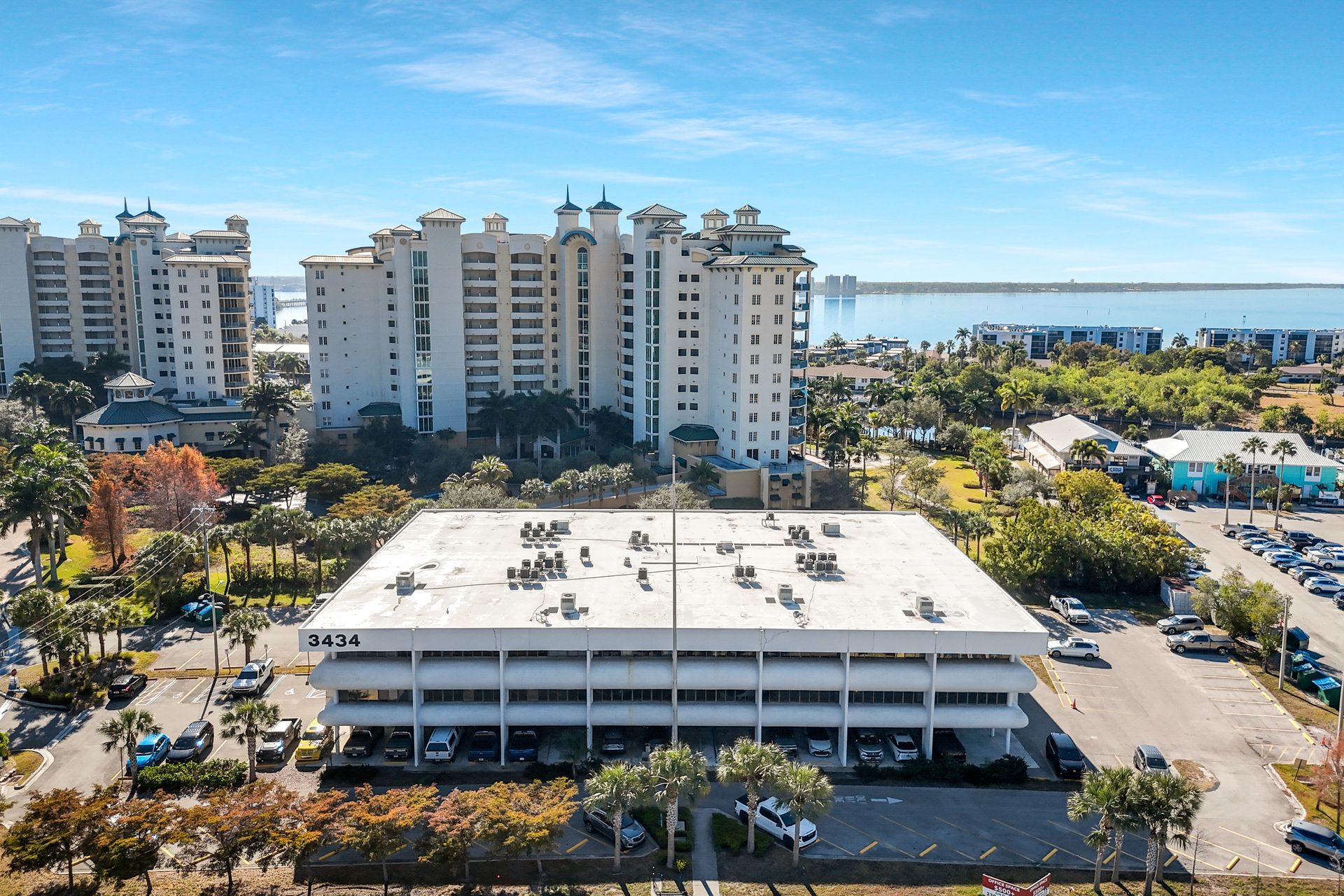 Aerial view of a three-story white office building with a flat roof, palm trees, and condos in the background; sunny day.