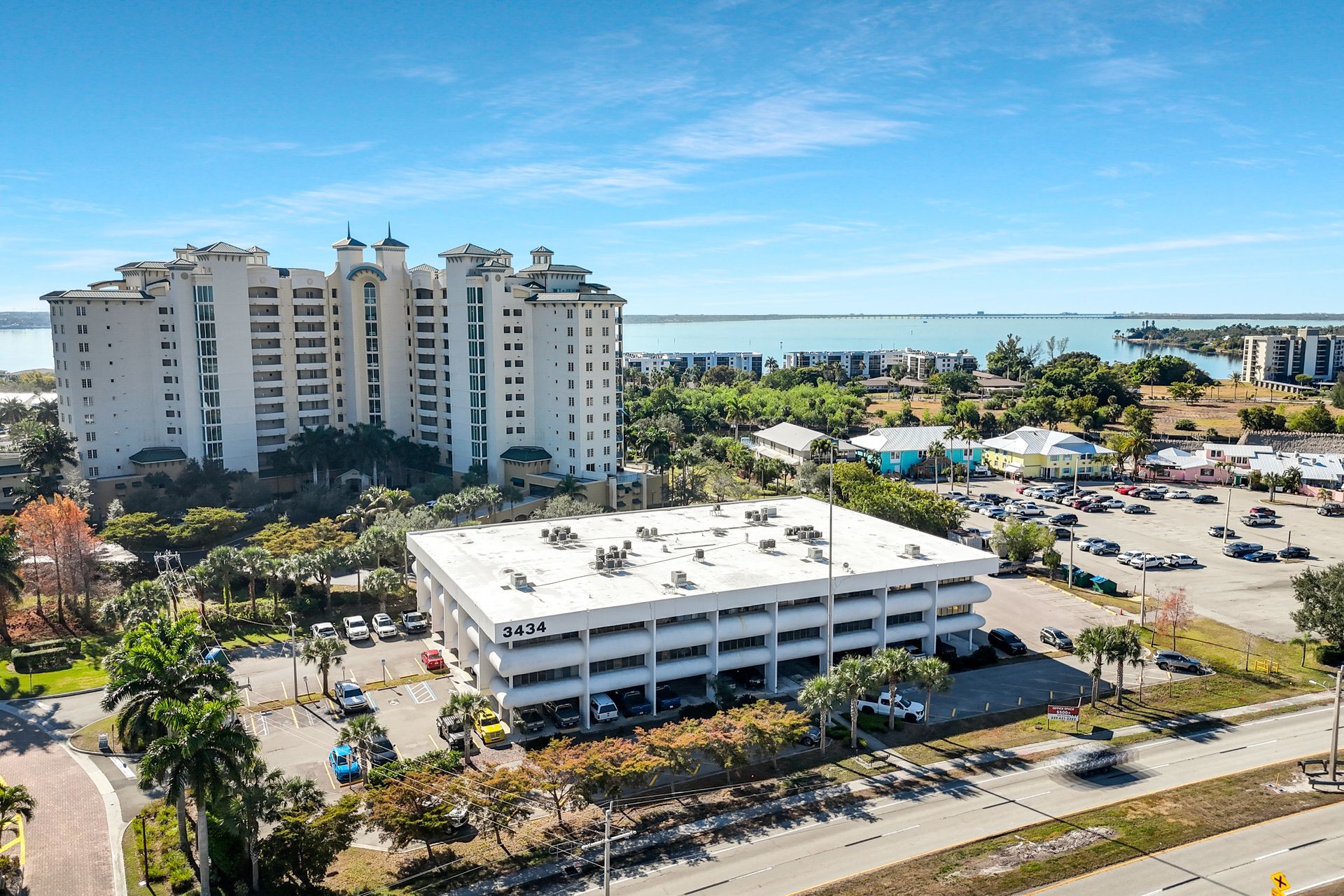 An aerial view of a three-story beige building with many parked cars in front and a large condo in the background, near the water.