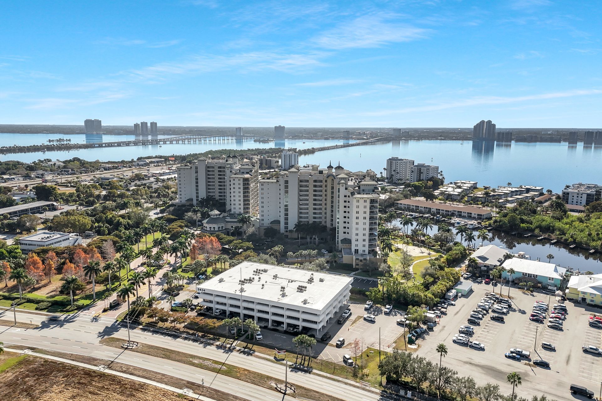 Aerial view of coastal buildings, parking lot, and water under a blue sky.