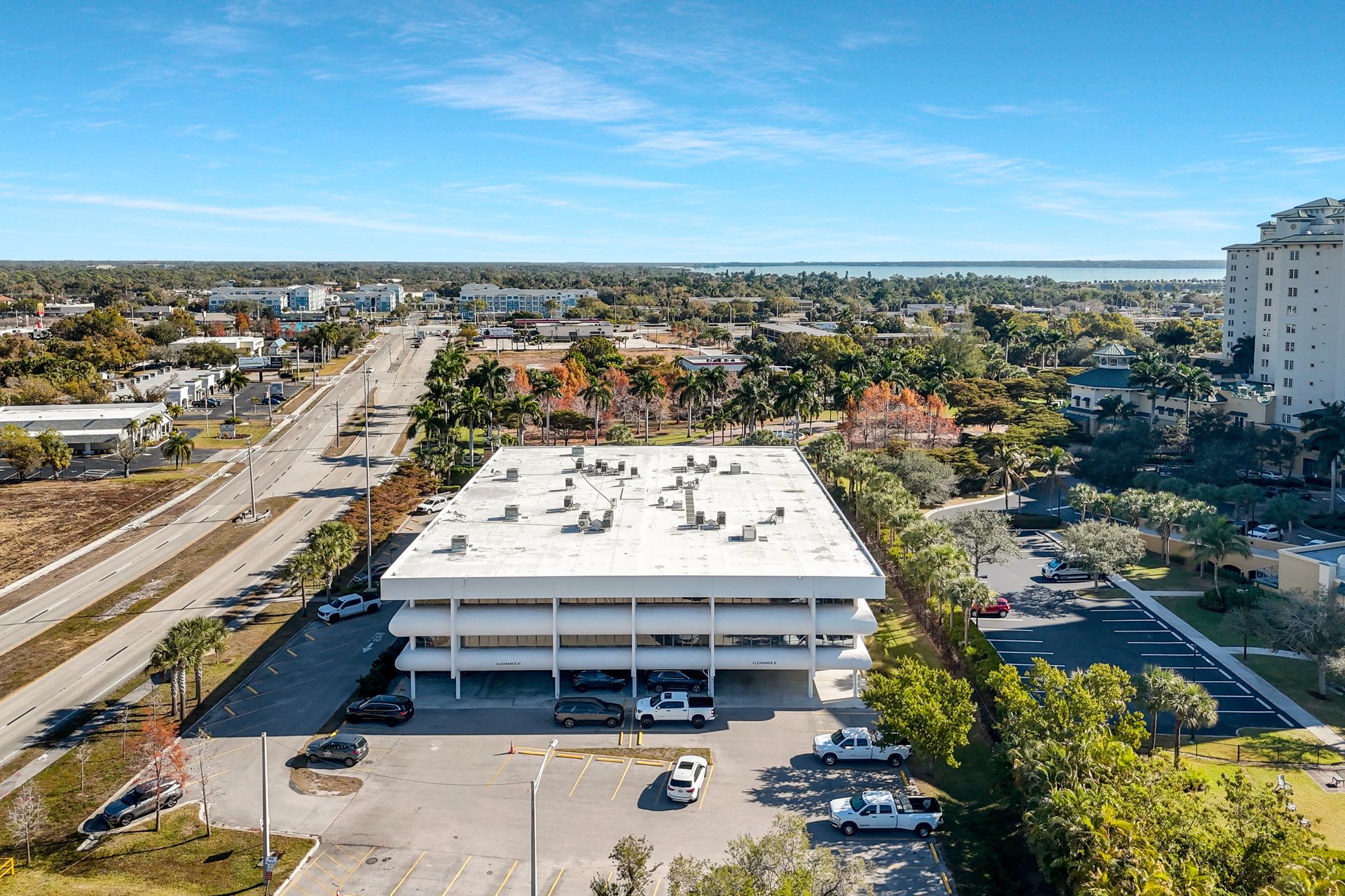 Aerial view of a white three-story commercial building with cars in a parking lot. Blue sky, cityscape background.