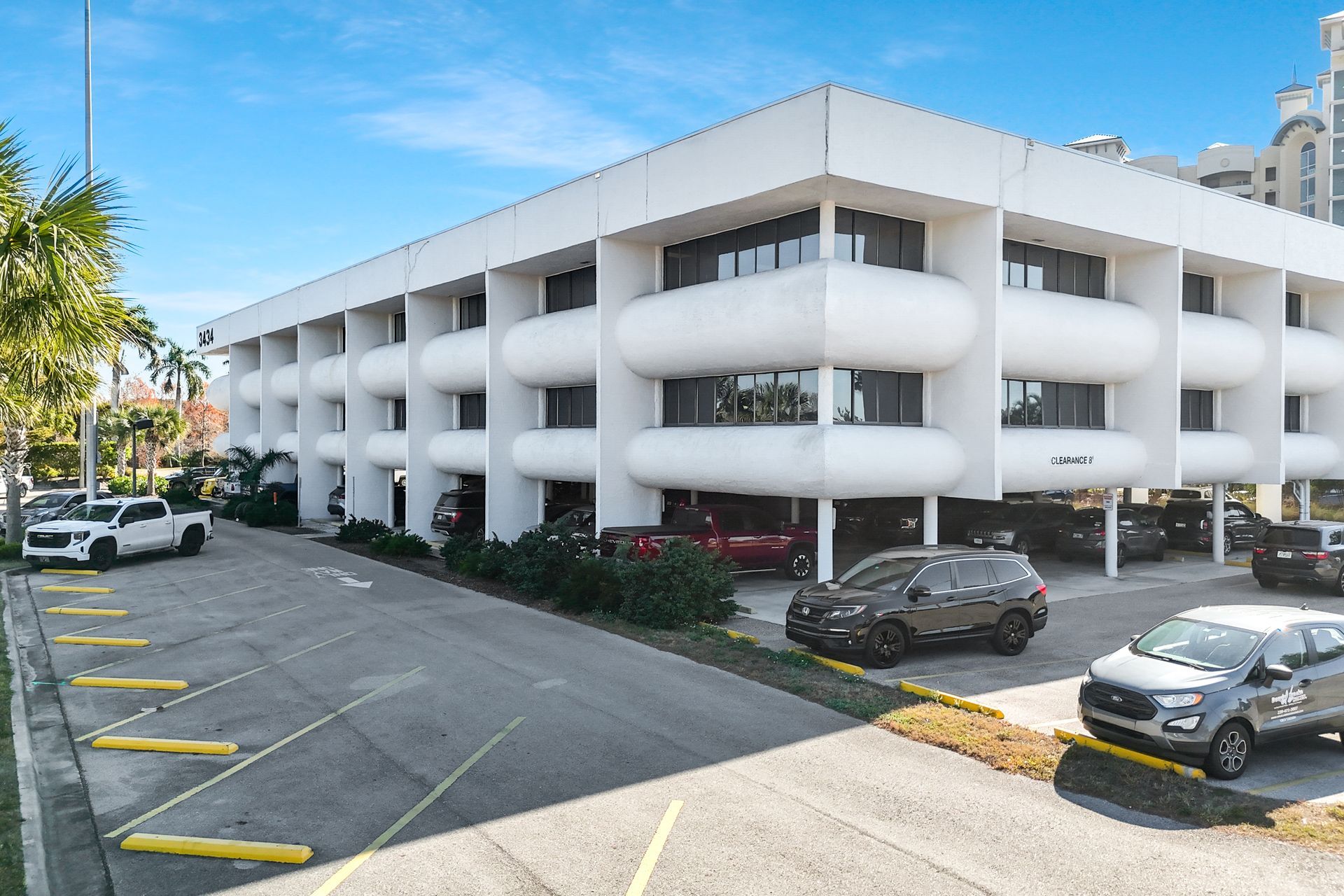 White office building with rounded balconies and parked cars in front.