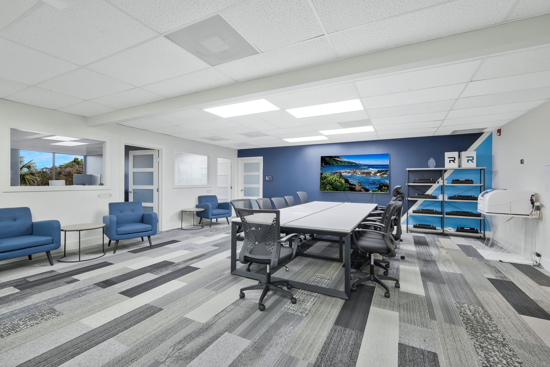 Meeting room with long table, chairs, blue accents, large screen showing scenery, and patterned carpet.