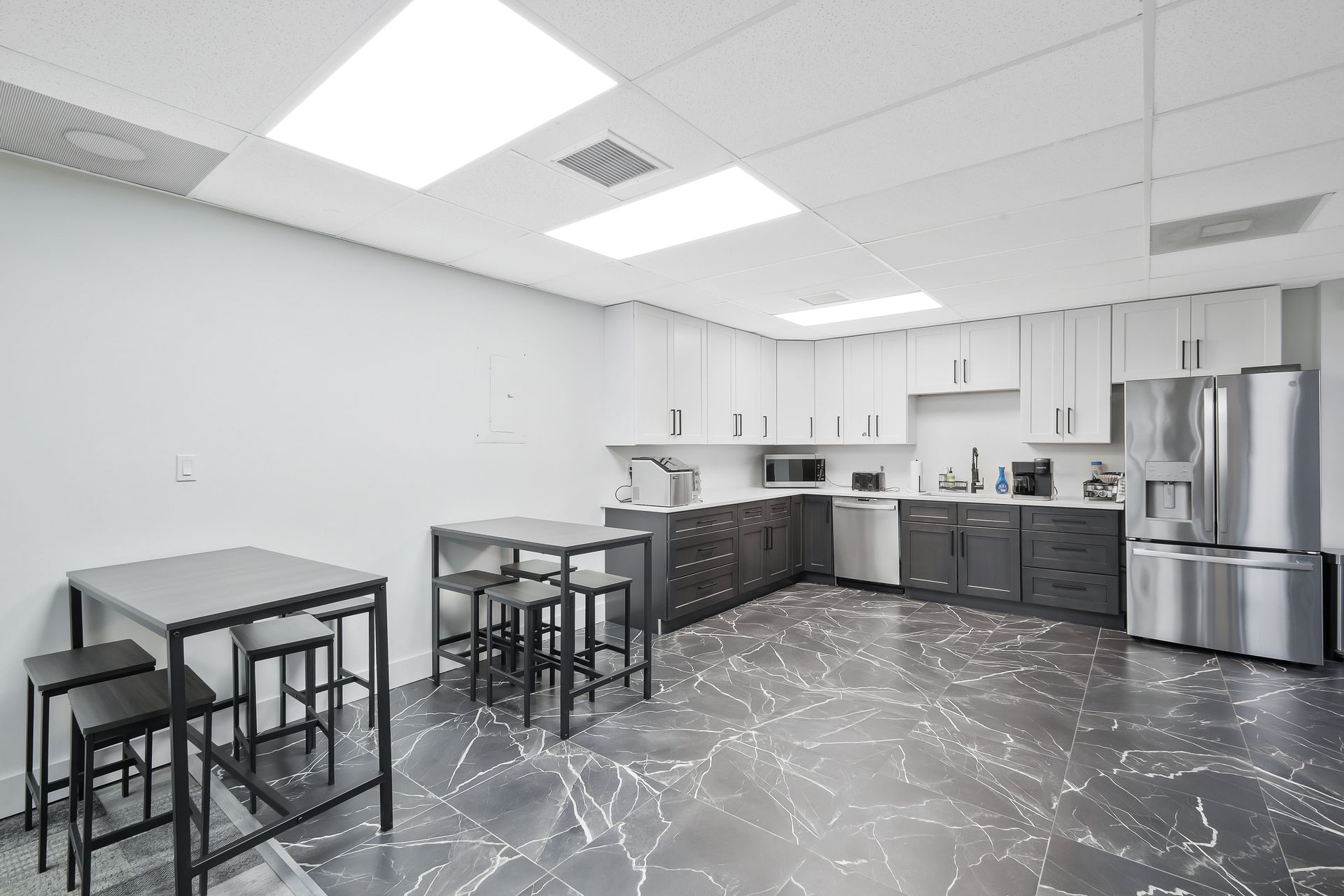 Modern office break room with kitchen cabinets, stainless steel refrigerator, and two tables with stools.