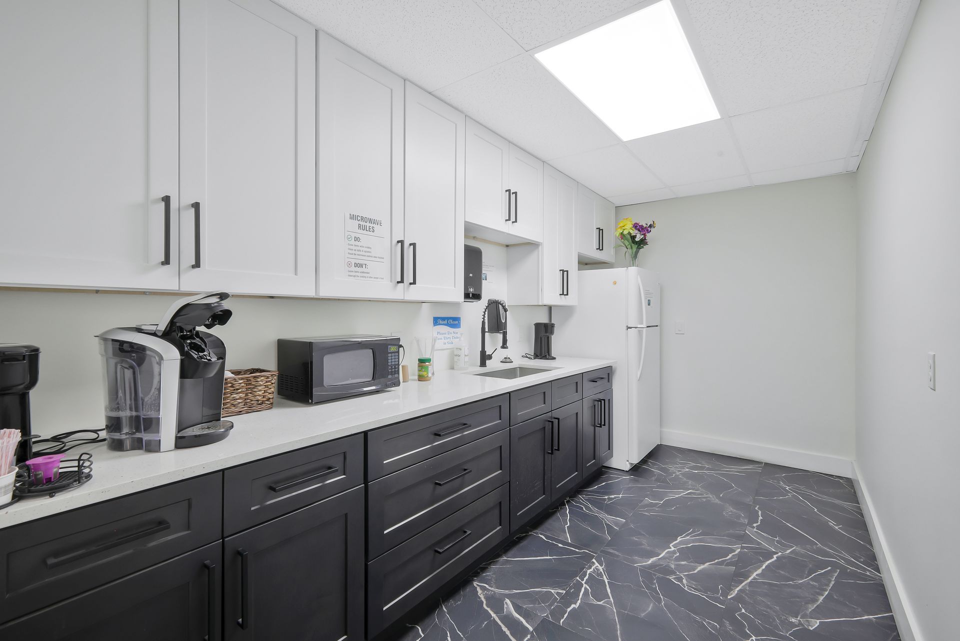 Office kitchenette with white and black cabinets, appliances, and patterned floor.