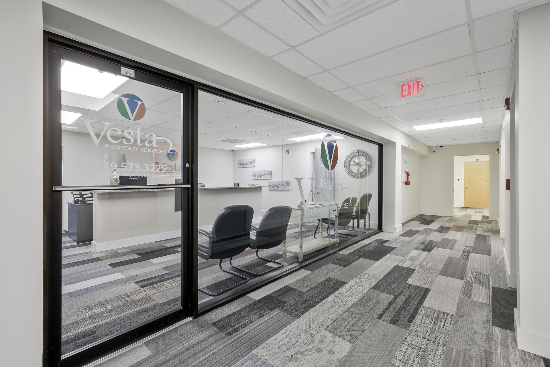 Office hallway with glass-walled waiting area. Vested logo on glass. Gray patterned carpet and white walls.