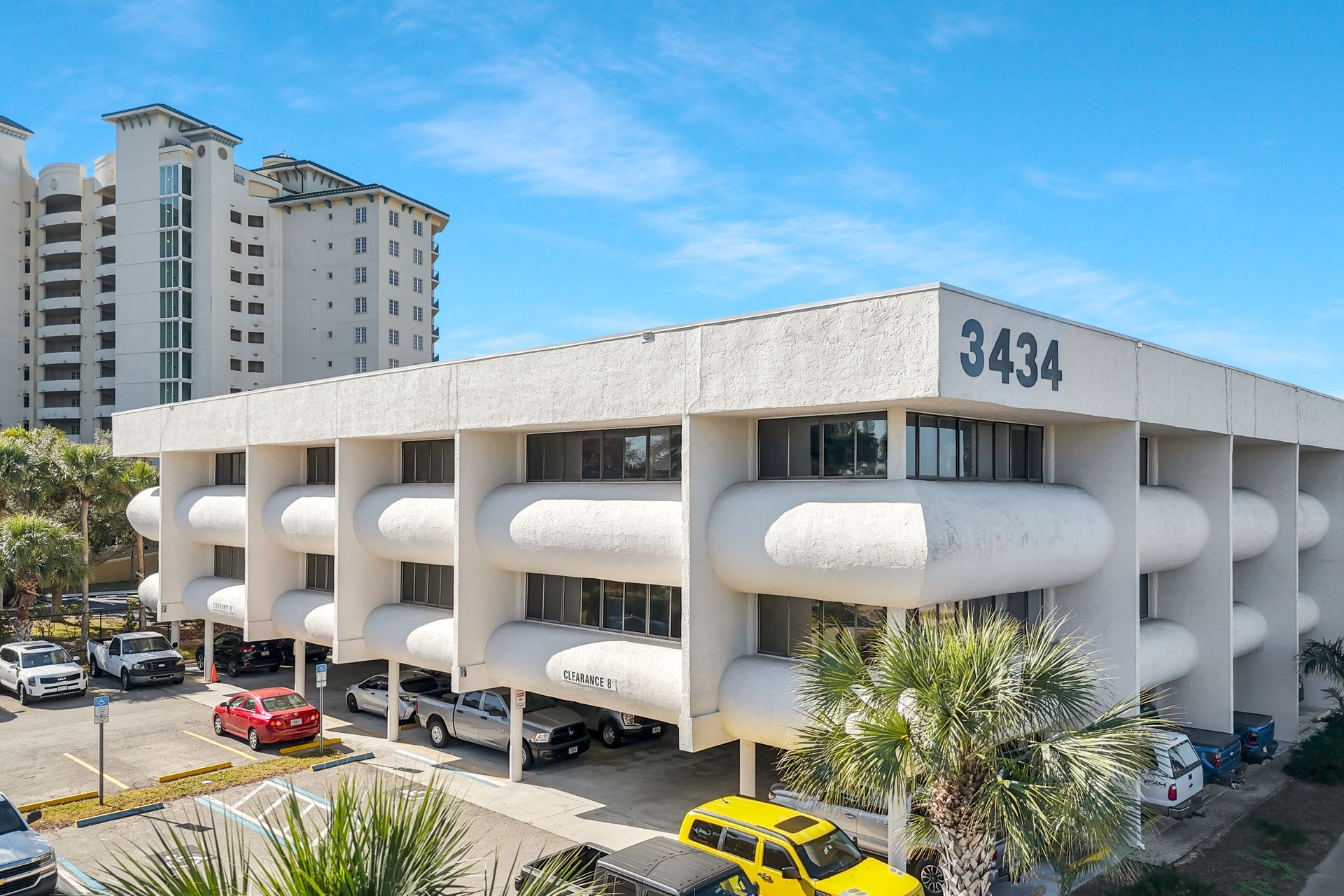 Three-story beige office building, number 3434, with cars parked below and a high-rise building in the background.