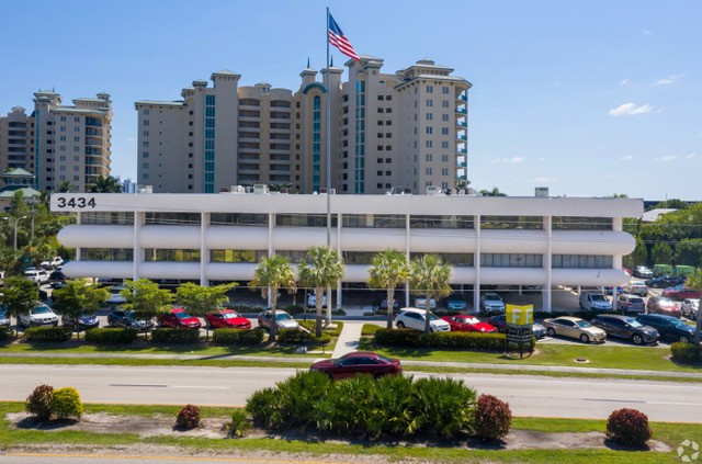 White office building with US flag, palm trees, cars, and a high-rise in the background.