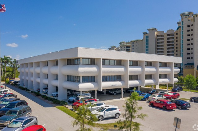 White, three-story office building with parking; American flag on left. Blue sky, cars parked outside.