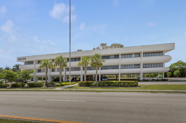 White office building with rounded edges, palm trees, and a road in front.