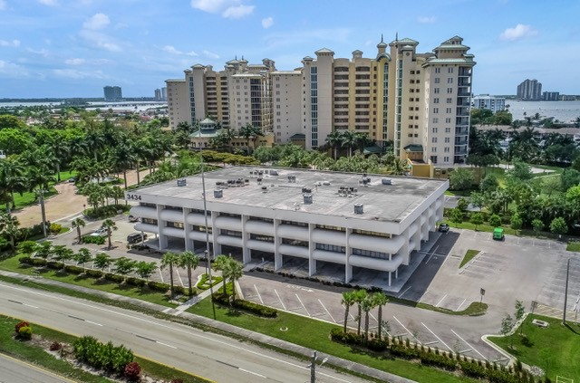 An aerial view of a three-story white commercial building with surrounding palm trees and large condo buildings.