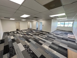Empty office space with patterned carpet and white walls. Fluorescent lights and windows visible.