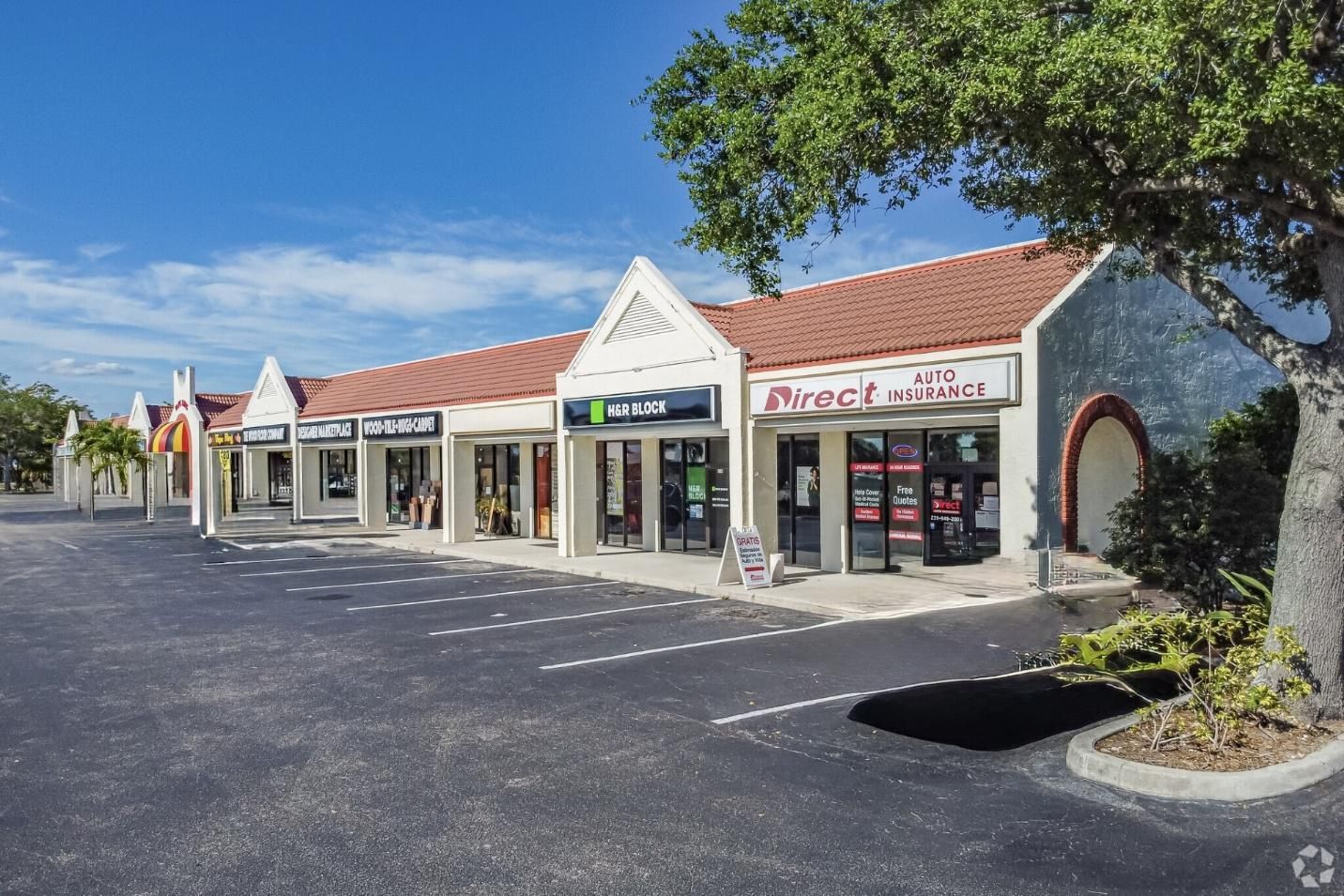 Shops in a strip mall with red tile roof, blue sky, and asphalt parking lot.