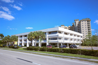 Three-story white office building with a condo building in the background. Blue sky and palm trees.