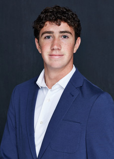 Young man in a blue blazer and white shirt smiles, against a dark background.