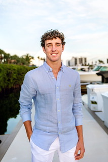 Young man with curly hair smiles, stands on a dock in blue shirt, white pants, hands in pockets.