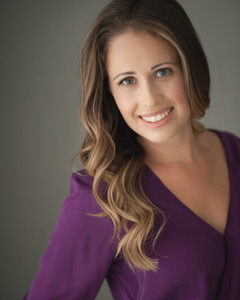 Woman with long wavy brown hair smiles, wearing a purple top, against a gray background.