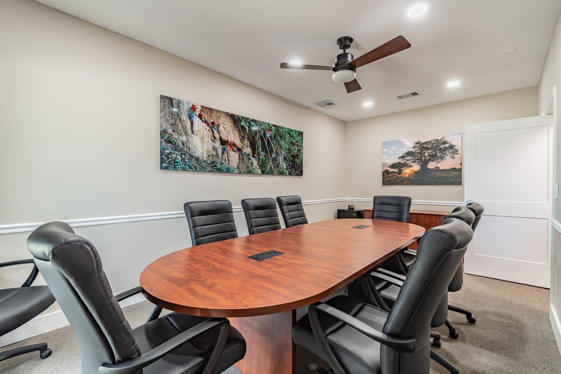 Conference room with oval wooden table, black leather chairs, and landscape art on wall.