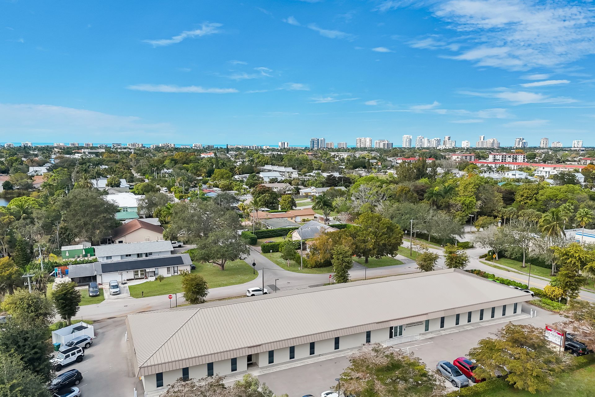 Aerial view of a suburban area with a long, low building in the foreground, trees, and buildings in the distance.