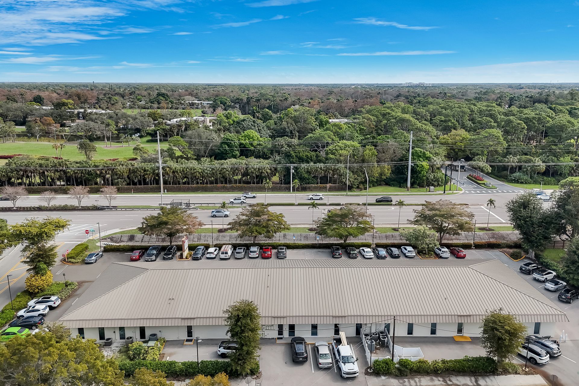 Aerial view of a single-story building with a flat roof, parking lot, and road, with trees and greenery in the background.