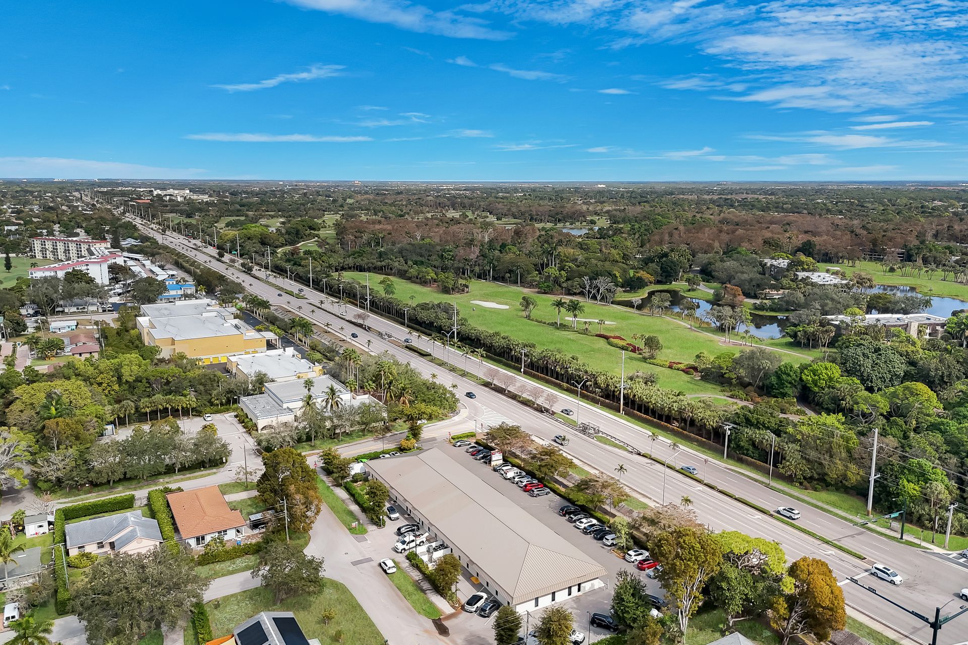 Aerial view of a long road lined with palm trees, buildings, and a golf course under a blue sky.