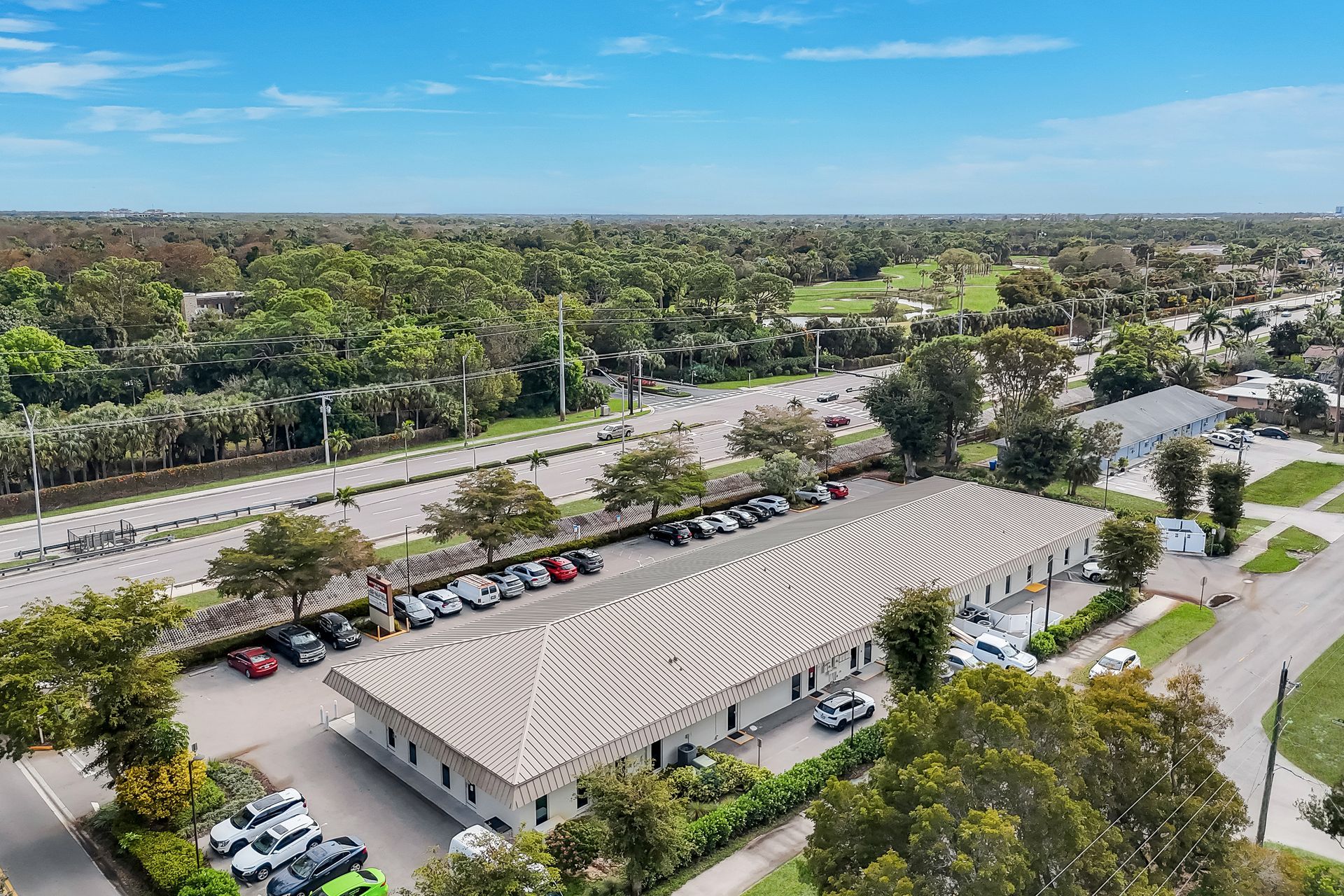 Aerial view of a long, white building with a patterned roof, cars parked outside, and a lush green landscape.