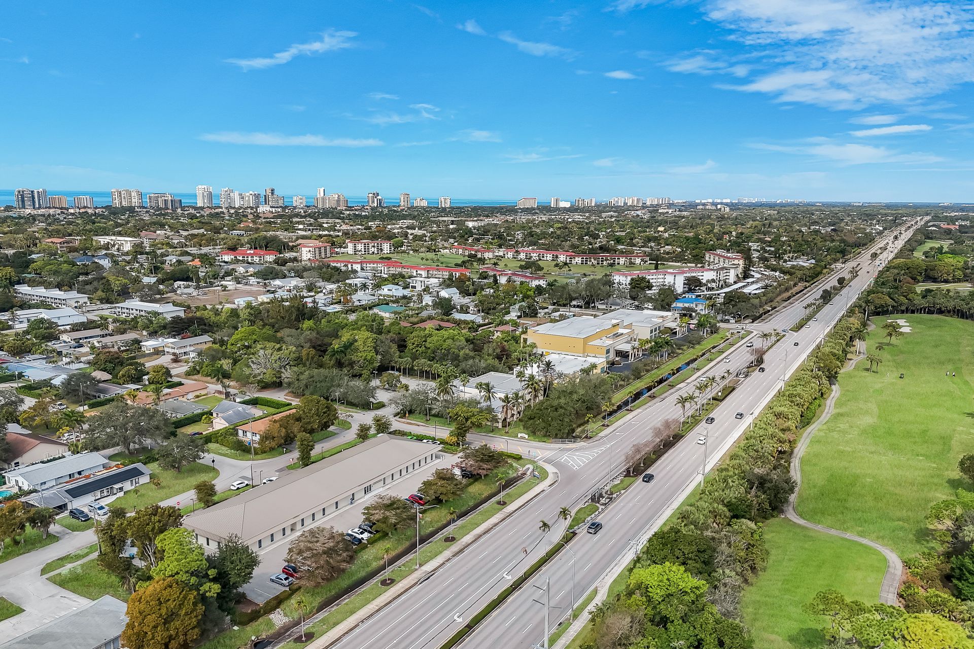 Aerial view of a city with a long road, buildings, and green space under a blue sky.