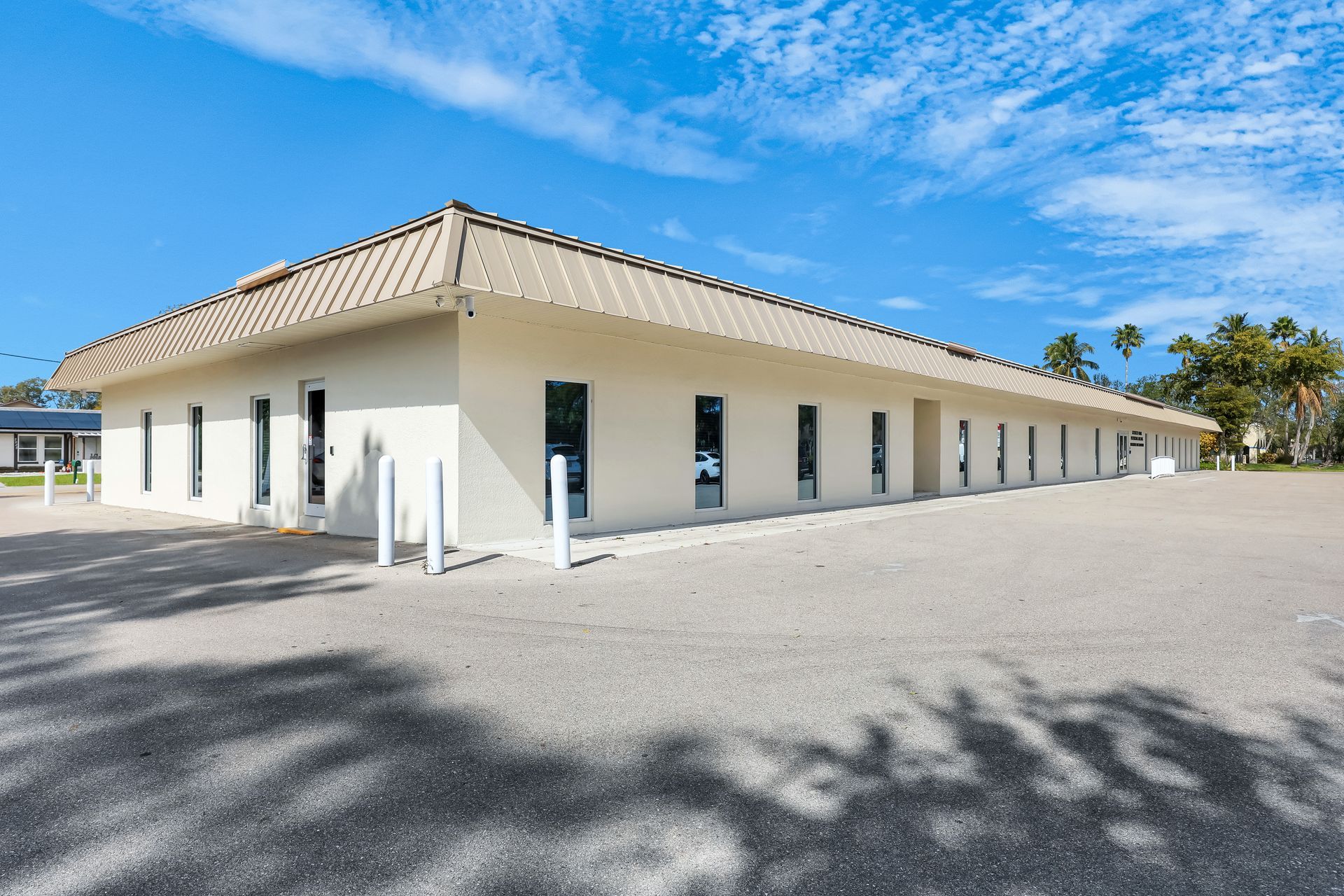 Single-story, beige commercial building with a tan roof. Parking lot in front, blue sky with clouds overhead.