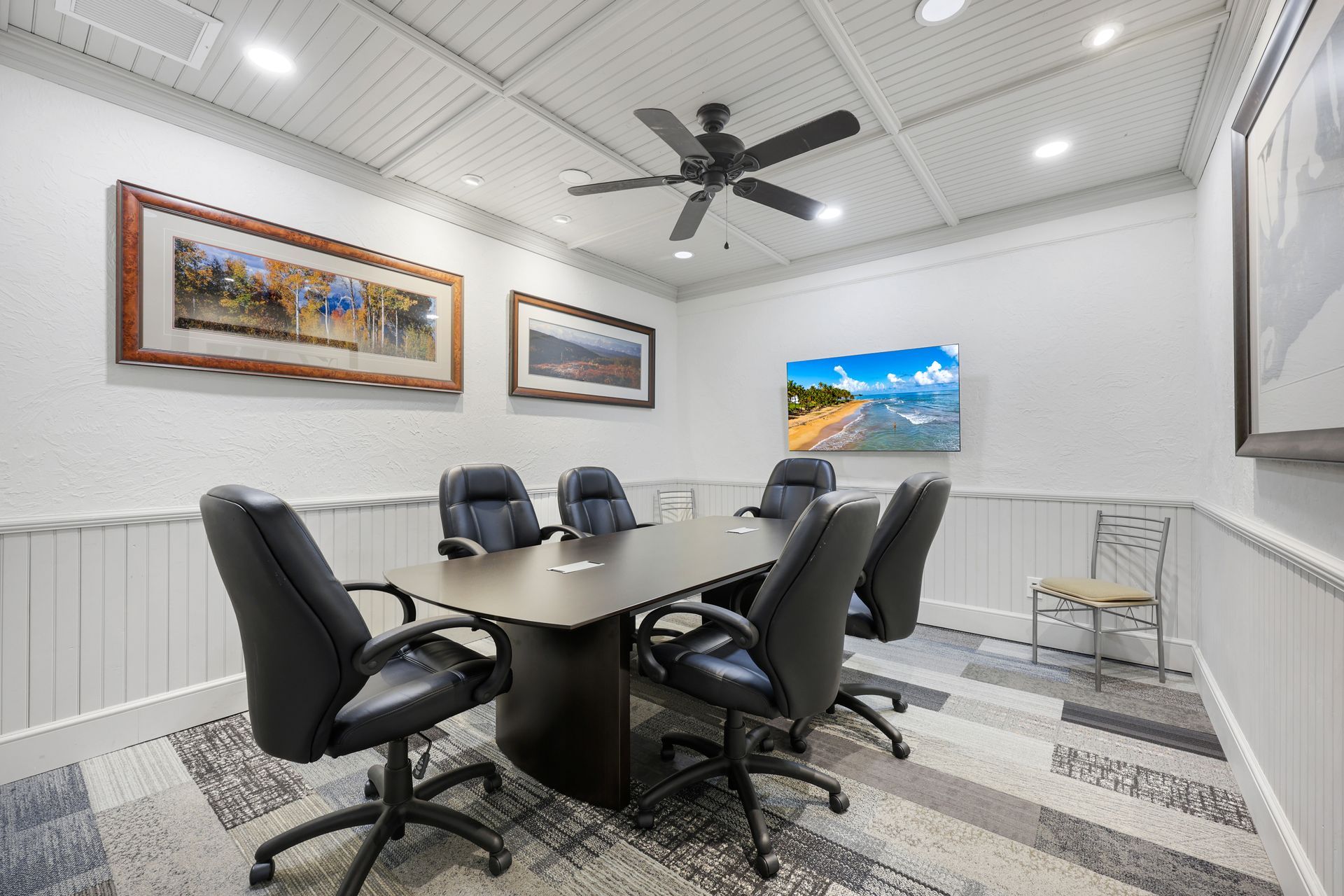 Conference room with a rectangular table, six black chairs, art on the walls, and a ceiling fan.