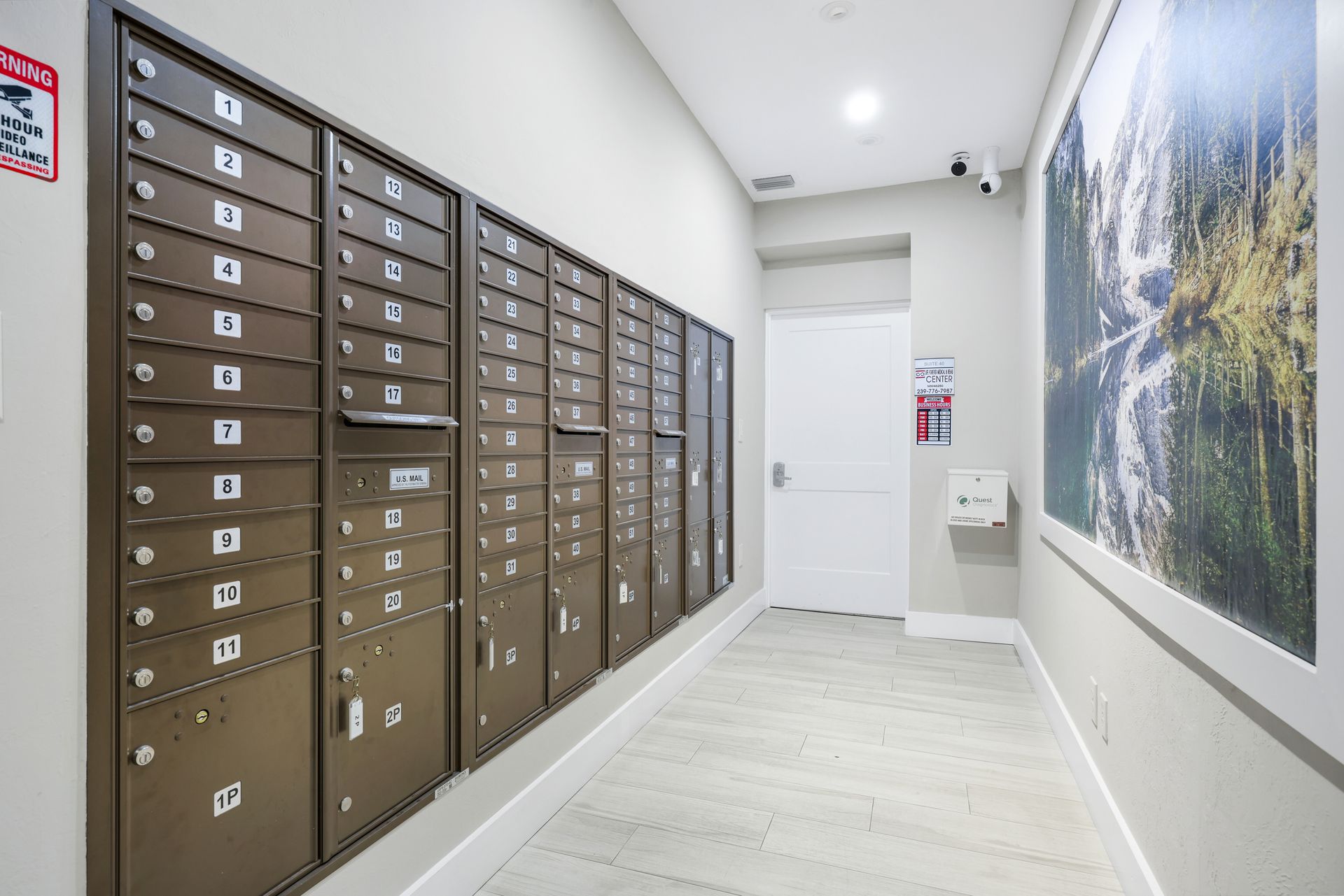 Mailboxes in a hallway, tan metal, beige walls, white door, and framed artwork on right.