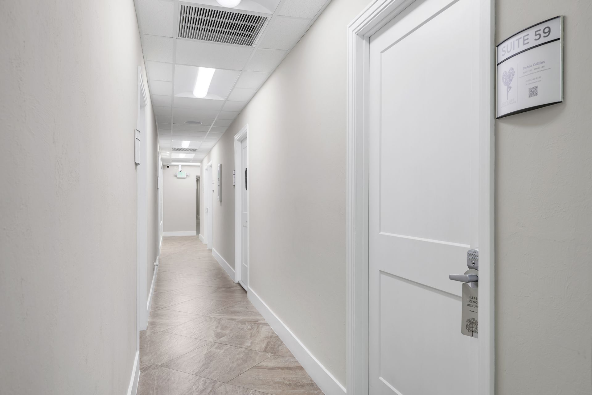 Hallway with white walls, doors, and a tile floor. Sign on door on the right.