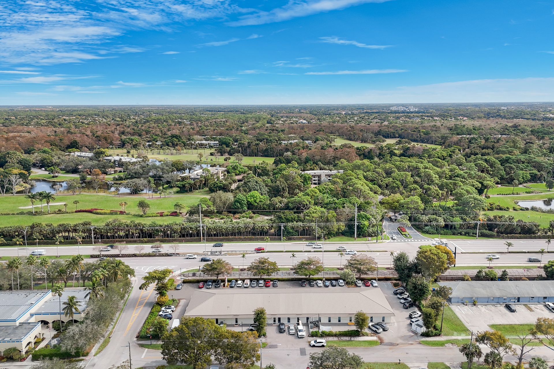 Aerial view of a commercial building with cars parked in front, with green trees and a road in the distance.