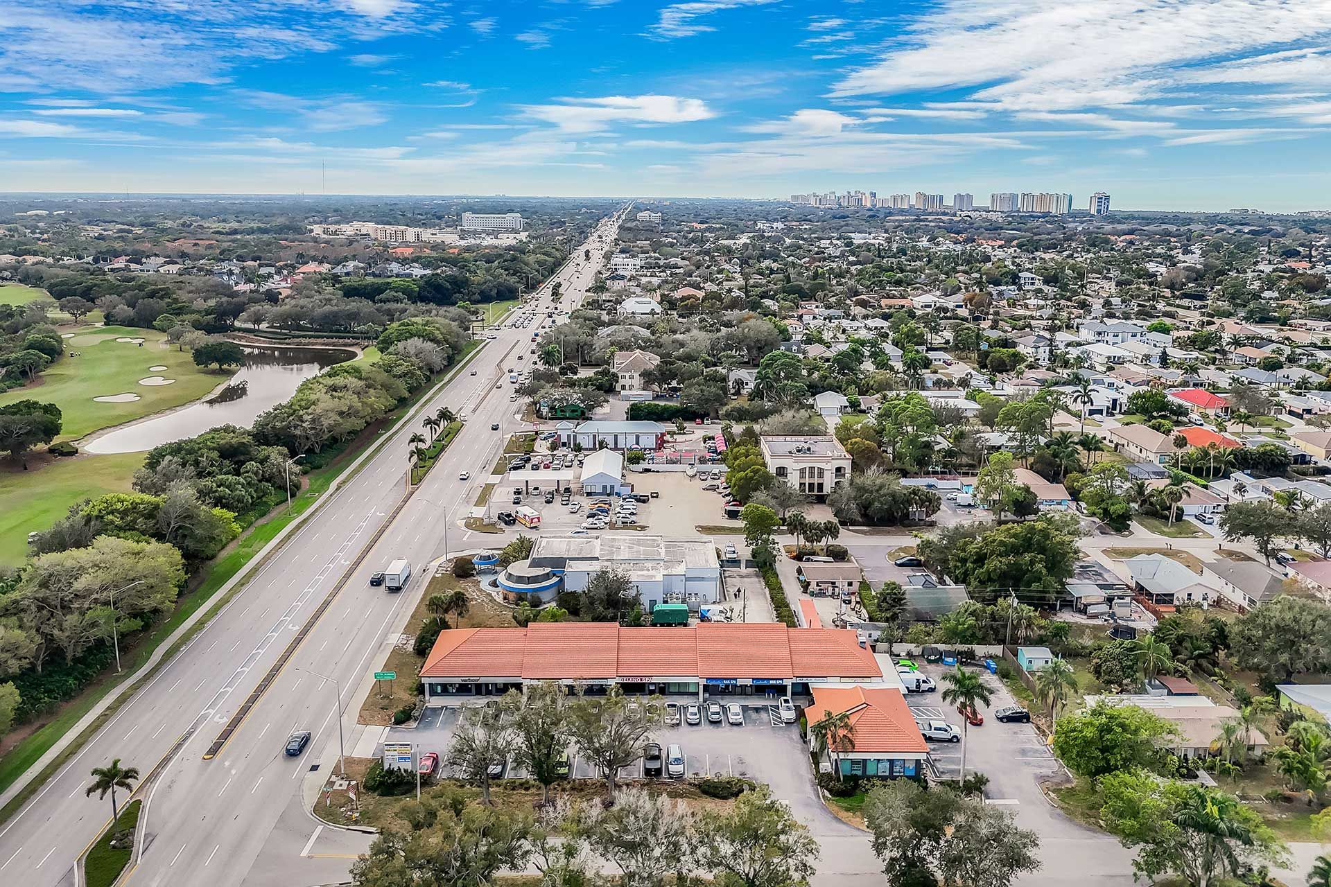 Aerial view of a long road lined with buildings and trees, a golf course on the left, and a distant city skyline.