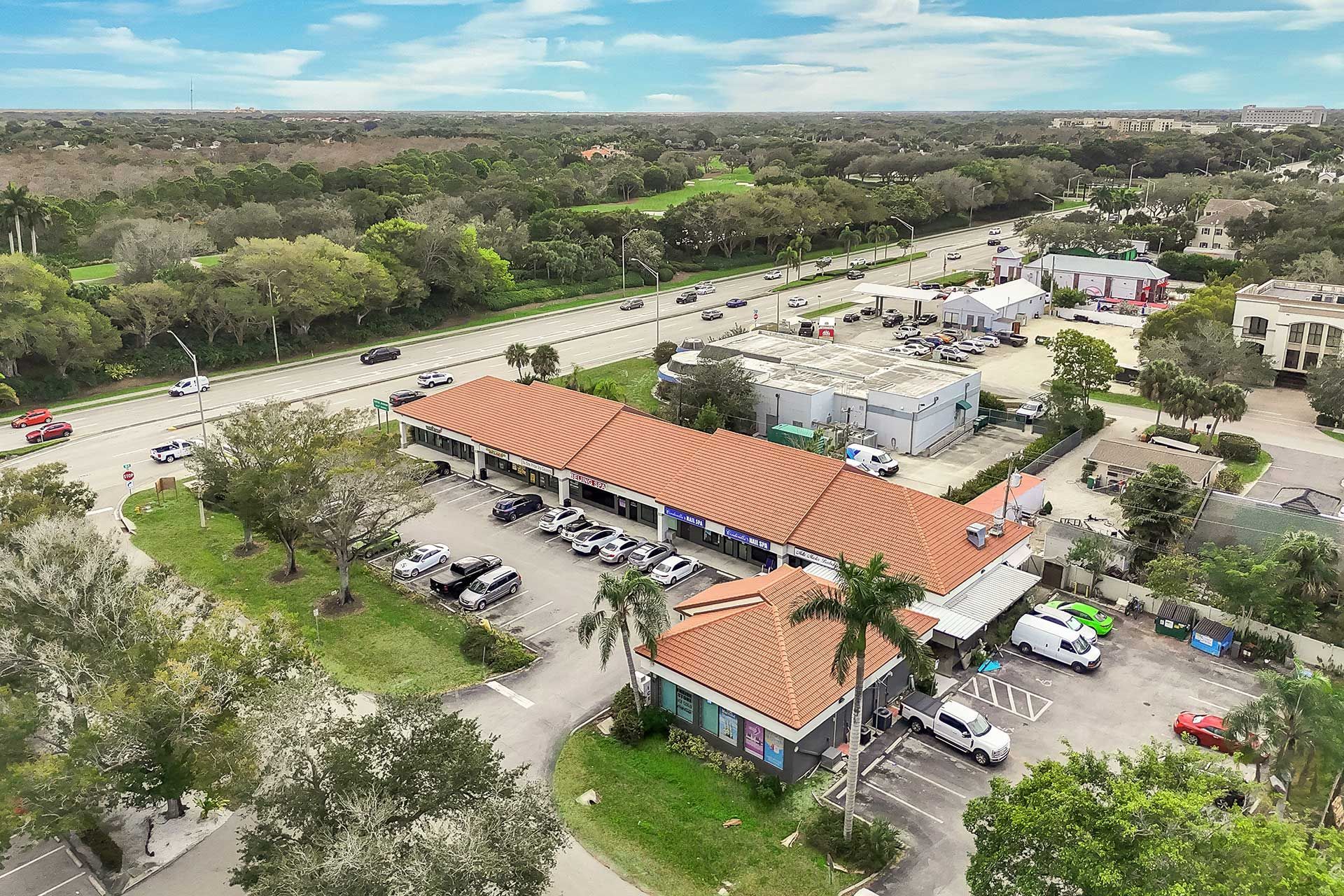 Aerial view of a commercial strip with a red tile roof, cars parked in front, and a busy road in the background.